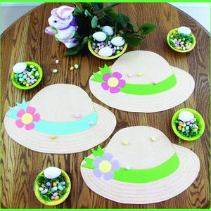 以下が含まれることがあります： 緑と青のリボンとピンク、パープル、イエローの花が付いた、日よけ帽子の形をしたベージュの布製のプレースマットが3つ。プレースマットは、キャンディーの卵が入ったボウルのある木製のテーブルの上に置かれています。