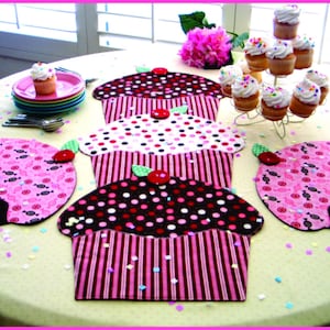 May include: A set of four pink and black cupcake-shaped table runners with white polka dots and stripes. The runners are arranged on a white tablecloth with confetti.