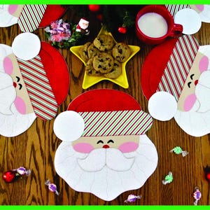 May include: Four red and white striped Santa Claus placemats with white felt faces and beards. The placemats are on a wooden table with chocolate chip cookies and sweets scattered around.