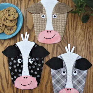 May include: Three cow-shaped pot holders with different fabric patterns. The pot holders are white with black and white spots, brown and white houndstooth, and grey and white plaid. Each pot holder has a pink nose, black eyes, and a pink smiling mouth.