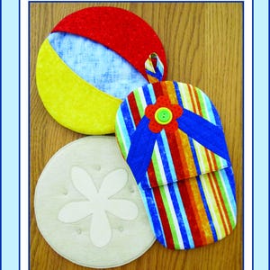 以下が含まれることがあります： ビーチをテーマにした布製の鍋つかみとマグカップマットが3つ。1つは赤と黄色のビーチボール、もう1つは青と赤のストライプのビーチサンダル、最後はベージュの貝殻です。