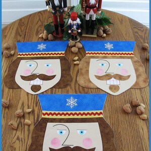 May include: Three nutcracker-themed placemats with embroidered faces and blue hats. The placemats are brown, beige, and blue. The placemats are on a wooden table with walnuts.