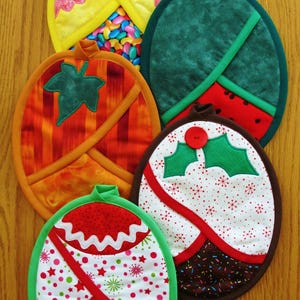 Puede incluir: Cinco agarraderas de olla acolchadas de colores con diferentes diseños. Las agarraderas de olla son de forma ovalada y tienen un borde verde, rojo y blanco. Los diseños incluyen un pollito, una sandía, una rama de acebo, un adorno navideño y una hoja.