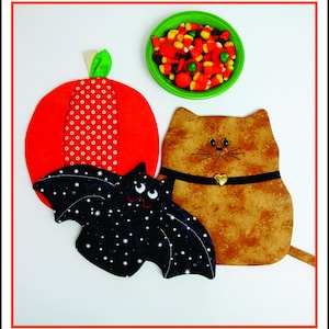 May include: A set of three Halloween-themed fabric pot holders. The pot holders are shaped like a pumpkin, a cat, and a bat. The pumpkin is red with white polka dots, the cat is brown with a black collar and a gold heart charm, and the bat is black with white stars.