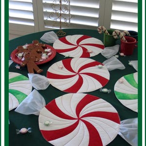 Op de afbeelding: Vier ronde placemats met een rode en witte pepermunt-spiraal met witte stoffen strikken op een groene tafelkleed. De placemats hebben een snoepstokdesign. Er liggen ook wat kleine groene en witte pepermuntjes op tafel.