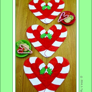 Puede incluir: Tres manteles individuales en forma de corazón rojo y blanco con forma de bastón de caramelo con detalles de acebo verde. Los manteles individuales están sobre una superficie de madera. Candy Cane Holidays Placemats, Table-Runner Or Table Topper. 2014, S. Tigue Shore. Susie C Shore Designs. A Division Of Tigue Productions, Inc.