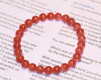 May include: A circular stretch bracelet composed of numerous polished, translucent red beads. The beads exhibit subtle variations in color intensity and internal texture, arranged in a continuous loop on a white surface with printed text.