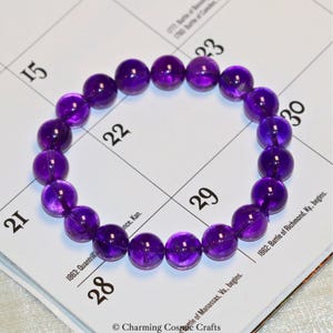 May include: A bracelet composed of spherical, polished, deep purple amethyst beads. The beads are strung together in a circular form. The bracelet is displayed on a calendar page with visible dates.