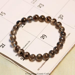 Puede incluir: Pulsera de cuentas de cuarzo ahumado translúcido, presentada sobre un calendario. Las cuentas tienen inclusiones internas y variaciones de color. Cada cuenta mide aproximadamente 0,8 cm de diámetro. Una joya.