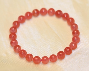 May include: A circular bracelet composed of small, faceted, translucent coral-pink gemstone beads strung together. The beads have a polished, reflective surface that catches the light, creating a uniform and continuous beaded loop design.