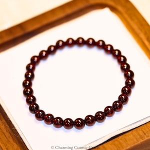 Puede incluir: Una pulsera de cuentas con piedras preciosas redondas y pulidas de color rojo oscuro. Las cuentas están ensartadas, formando un círculo completo. La pulsera se muestra sobre una superficie blanca dentro de una bandeja de madera. La imagen está bien iluminada.