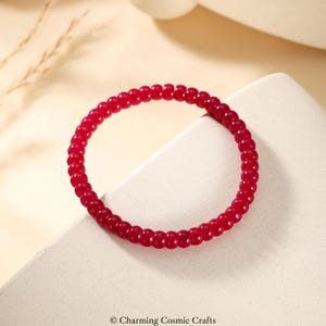 May include: A vibrant red beaded bracelet. The bracelet is composed of small, round beads, forming a continuous loop. The beads have a glossy finish, reflecting light. The bracelet is displayed on a neutral-toned surface.