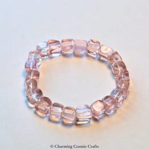 May include: A bracelet composed of light pink, square beads. The beads are translucent and strung together. The bracelet is displayed on a white surface. The text "Charming Cosmic Crafts" is visible in the bottom right corner.