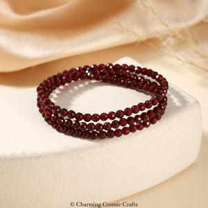 Könnte beinhalten: Dreireihiges Armband aus kleinen, runden, tiefroten Perlen. Die Perlen sind zu einem flexiblen Band aufgereiht. Das Armband wird auf einer weißen Oberfläche mit einem weichen, neutralen Hintergrund präsentiert.