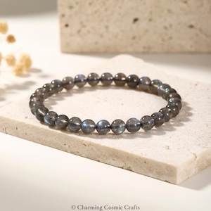 May include: A beaded bracelet made of dark gray labradorite stones. The round beads have iridescent blue and gray flashes. The bracelet is displayed on a light-colored, textured surface. The image is well-lit, highlighting the bracelet's details.