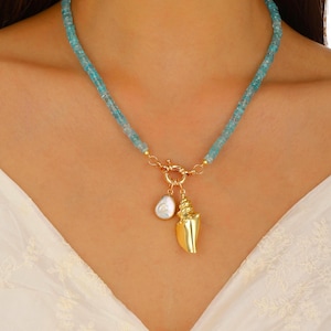 May include: A necklace featuring a strand of small, light blue cylindrical beads. The necklace includes a gold-toned circular clasp at the center, from which hang two charms: one irregular white pearl and one gold-toned seashell pendant.