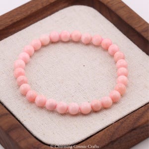 Peut inclure: Un bracelet de perles rose clair. Le bracelet est composé de perles rondes et polies. Les perles sont enfilées sur un élastique. Le bracelet est présenté sur un tissu beige à l'intérieur d'un plateau en bois. C'est un bijou délicat.