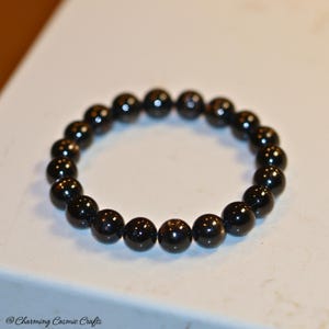 Puede incluir: Una pulsera elástica circular compuesta por cuentas negras lisas y pulidas dispuestas en un bucle continuo. El texto "@ Charming Cosmic Crafts" está impreso en la esquina inferior izquierda del marco de la imagen.