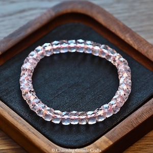 May include: A pink gemstone bracelet with cut beads. The bracelet is presented on a black surface inside a wooden tray. The beads are a soft pink colour, reflecting light. The bracelet is a piece of jewellery.
