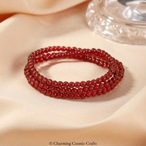 Könnte beinhalten: Armband aus drei Strängen roter Edelsteinperlen. Die Perlen sind klein und rund, mit einer tiefroten Farbe. Das Armband wird auf einem cremefarbenen Stoff präsentiert. Das Bild ist gut beleuchtet und zeigt die Details.
