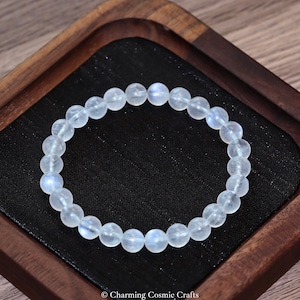 Op de afbeelding: Een armband met heldere, ronde kralen met een subtiele blauwe tint. De armband ligt op een zwarte ondergrond in een houten bakje. De tekst "Charming Cosmic Crafts" is onderaan zichtbaar.