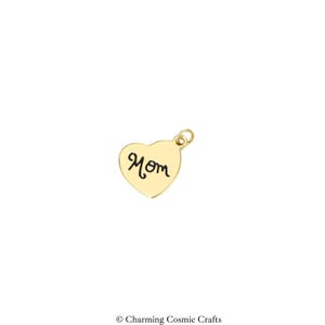 May include: A gold-toned heart-shaped charm featuring the word Mom engraved in a black script font. The charm includes a small circular jump ring at the top for attachment, presenting a smooth, polished surface finish on the metal heart pendant.