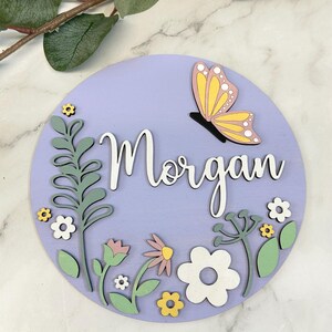 May include: A round, lavender wooden sign with the name "Morgan" in white script. A yellow and pink butterfly is perched above the name. The sign is decorated with green, white, and yellow floral elements.
