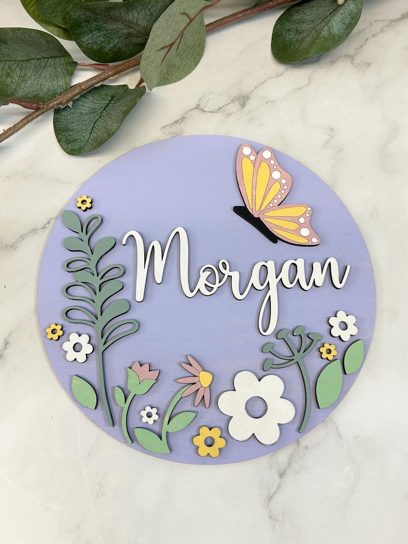 May include: A round, lavender wooden sign with the name "Morgan" in white script. A yellow and pink butterfly is perched above the name. The sign is decorated with green, white, and yellow floral elements.