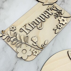 May include: Wooden sign with the name "Kianna" in a decorative font, surrounded by laser-cut floral and butterfly accents. The sign is made of light-colored wood and features a rectangular shape with rounded corners. The butterfly is at the top.