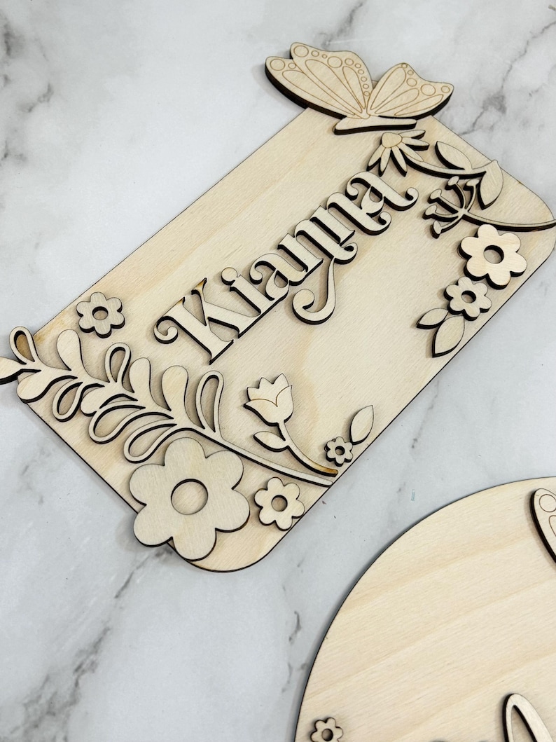 May include: Wooden sign with the name "Kianna" in a decorative font, surrounded by laser-cut floral and butterfly accents. The sign is made of light-colored wood and features a rectangular shape with rounded corners. The butterfly is at the top.