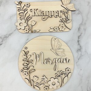 May include: Two wooden name signs. One is rectangular with the name "Kianna" and floral and butterfly accents. The other is round with the name "Morgan" and similar floral and butterfly details. Both are made of light-colored wood.