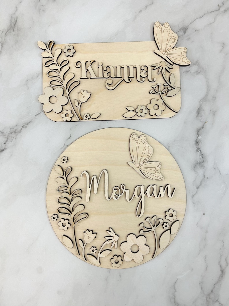 May include: Two wooden name signs. One is rectangular with the name "Kianna" and floral and butterfly accents. The other is round with the name "Morgan" and similar floral and butterfly details. Both are made of light-colored wood.