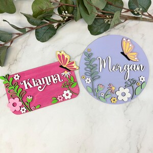May include: Two wooden name signs. One is rectangular and pink with the name "Kianna" and floral accents. The other is round and lavender with the name "Morgan" and floral accents. Both signs feature butterfly embellishments.