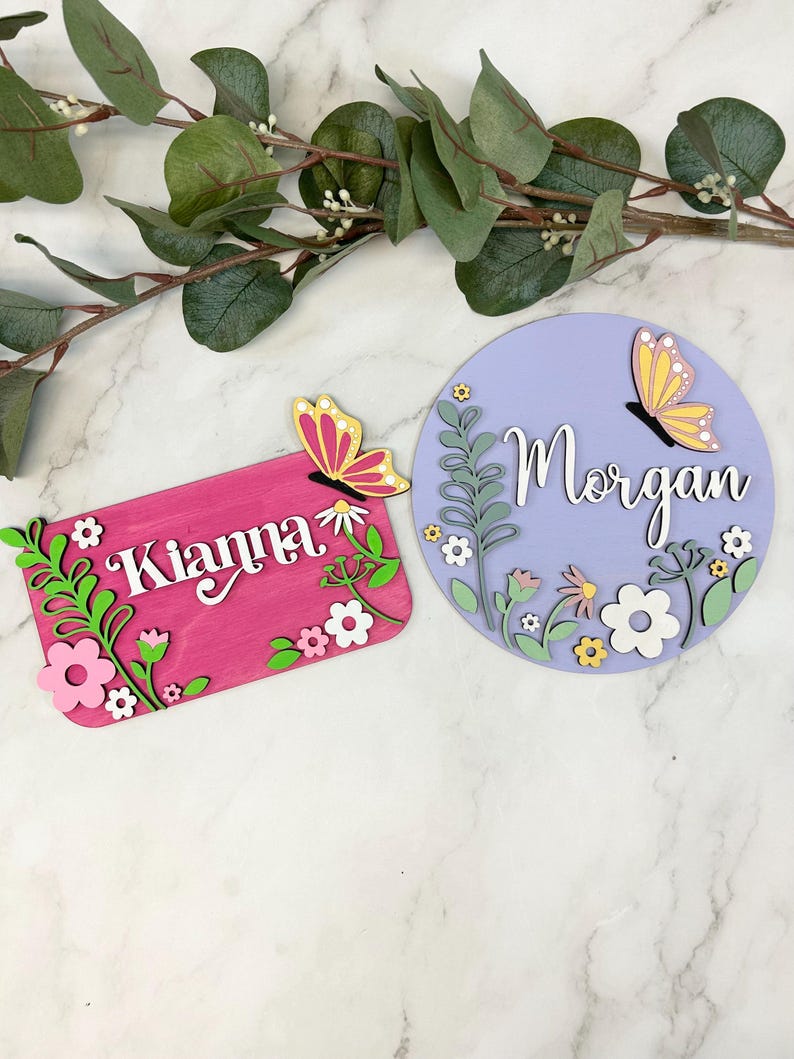 May include: Two wooden name signs. One is rectangular and pink with the name "Kianna" and floral accents. The other is round and lavender with the name "Morgan" and floral accents. Both signs feature butterfly embellishments.