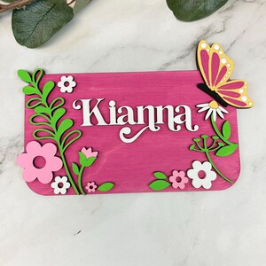 May include: A pink wooden sign with the name "Kianna" in white script. The sign is decorated with green leaves, pink and white flowers, and a butterfly with yellow and pink wings. The sign is likely a decorative item.