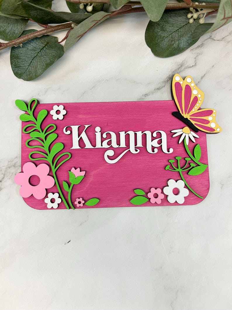 May include: A pink wooden sign with the name "Kianna" in white script. The sign is decorated with green leaves, pink and white flowers, and a butterfly with yellow and pink wings. The sign is likely a decorative item.