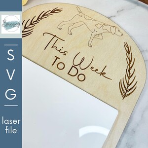 Puede incluir: Un letrero de madera con una superficie blanca de borrado en seco. El letrero tiene forma de semicírculo y tiene el texto "This Week To Do" escrito en él. Hay una pequeña ilustración de un perro en el letrero.