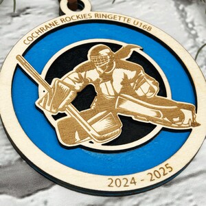 May include: A wooden hockey ornament with a blue and black background. The ornament features a hockey player in a goalie stance with a stick and a puck. The text "Cochrane Rockies Ringette U16B 2024-2025" is printed on the ornament.