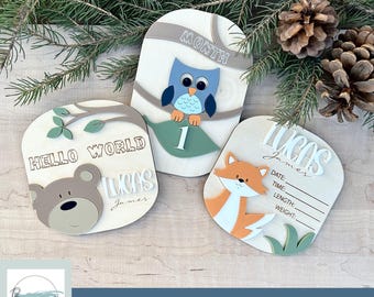 Puede incluir: Tres placas de madera para hitos de bebé con un tema de bosque. La primera placa presenta un oso marrón y el texto "Hello World Lucas James". La segunda placa presenta un búho y el texto "Mes 1". La tercera placa presenta un zorro y el texto "Lucas James" con líneas para la fecha, la hora, la longitud y el peso.