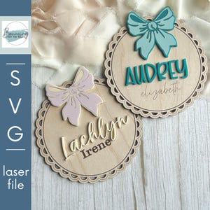 Può includere: Due targhette in legno tagliate al laser con bordi smerlati e dettagli a fiocco. Un'etichetta presenta un fiocco rosa chiaro e il nome "Lachlyn" con "Irene" sotto. L'altra ha un fiocco verde acqua e il nome "Audrey" con "elizabeth" sotto.
