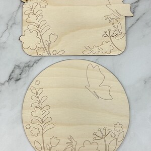 May include: Two unfinished wooden craft shapes: a rectangle and a circle. Both are decorated with etched floral and butterfly designs. The rectangle has a butterfly cutout on one corner. The circle features a butterfly and floral border.