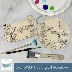 May include: Wooden laser-cut name signs with floral and butterfly designs. One sign is rectangular with the name "Kianna", the other is round with the name "Morgan". Paintbrushes and a paint palette with various colors are also visible. The text "SVG LASER FILE digital download" is at the bottom.