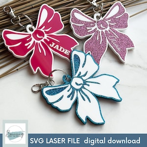 Op de afbeelding: Drie verschillende sleutelhangers in de vorm van een strik met glitter en witte contouren. De sleutelhangers zijn roze, blauw en rood. De roze sleutelhanger heeft de naam "Olivia" erop, de rode sleutelhanger heeft de naam "Jade" erop en de blauwe sleutelhanger heeft geen tekst.