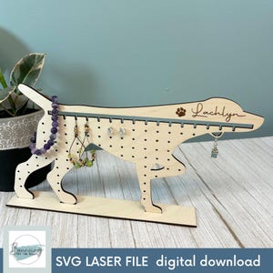Pode incluir: Um suporte de brincos de madeira em forma de cão com uma pegada de pata e o nome "Lachlyn" gravado na parte superior. O suporte tem vários orifícios para brincos e um espaço para uma pulseira.