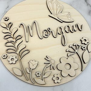 May include: A round wooden sign with the name "Morgan" in cursive script, surrounded by laser-cut floral and butterfly designs. The sign is made of light-colored wood, with intricate details in the cut-out elements.