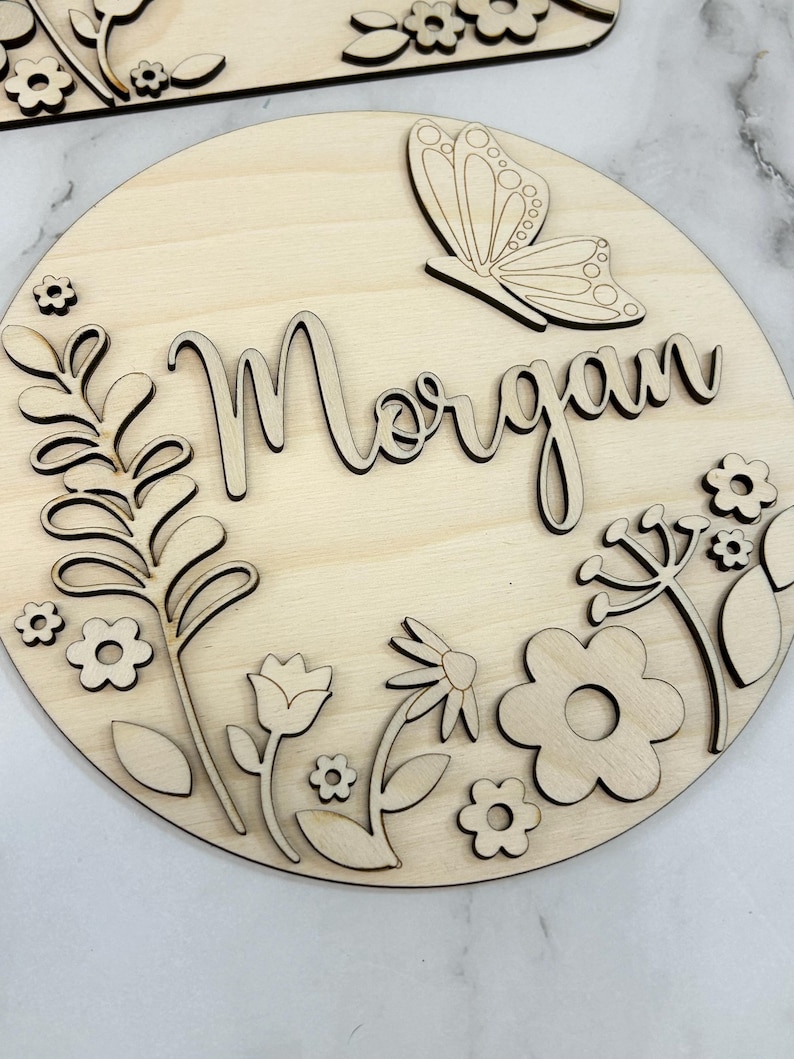 May include: A round wooden sign with the name "Morgan" in cursive script, surrounded by laser-cut floral and butterfly designs. The sign is made of light-colored wood, with intricate details in the cut-out elements.