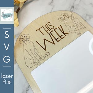 Puede incluir: Un letrero de madera con el texto "This Week" y dos ilustraciones de perros. El letrero tiene un espacio en blanco para escribir.