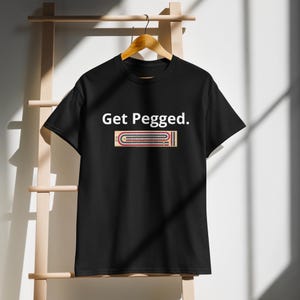 Könnte beinhalten: Schwarzes T-Shirt mit dem Text "Get Pegged." und einer Grafik eines Brettspiels mit Stiften.