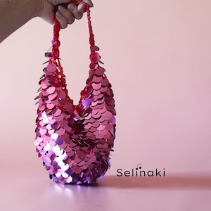 Puede incluir: Un bolso pequeño cubierto de lentejuelas metálicas de color rosa brillante superpuestas. El bolso cuenta con un asa roja de ganchillo y una forma profunda y redondeada. El texto "Selinaki" con una nota musical está impreso en negro abajo.