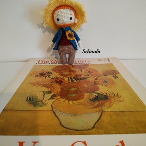Könnte beinhalten: Eine gehäkelte Puppe mit einem gelben Strohhut und einer blauen Jacke steht auf einem Buch mit dem Titel "Die großen Künstler" mit einem Gemälde von Sonnenblumen von Van Gogh auf dem Cover.